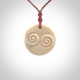 A hand carved and intricate koru pendant made for us by Yuri Terenyi. This is a beautiful little piece and is emblematic of the well known and loved Koru design. It is carved from bone in a round shape with decorative design carved into the koru. It is suspended from a red/khaki cord with a red floret and the necklace is adjustable.